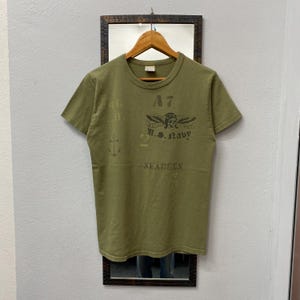 May include: Olive green t-shirt with a crew neck and short sleeves. The shirt features a distressed graphic print with the text "A7", "U.S. Navy", and "SEABEES", along with an anchor symbol. The shirt is hanging on a wooden hanger.