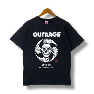 May include: Black t-shirt with the word "OUTRAGE" in white block letters. The shirt features a white skull graphic inside a circular design. Japanese text and a red stamp are below the graphic.