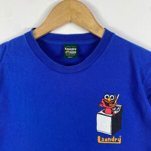 May include: A blue t-shirt featuring a graphic of a red character in a washing machine. The text "laundry" is printed in yellow at the bottom of the shirt. The shirt has a tag that reads "Laundry Japan" and "Small".