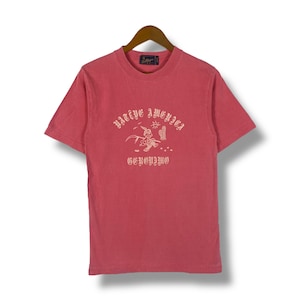 Peut inclure: Un t-shirt rose avec un imprimé graphique d'un personnage de dessin animé courant avec une faux. Le texte "WATT EYE AMERICA" et "GCKOUJIWO" est imprimé en police gothique.