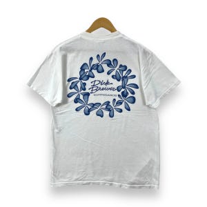 Puede incluir: Camiseta blanca con un diseño floral azul y el texto "Dick Brewer Surfboards" en la espalda.