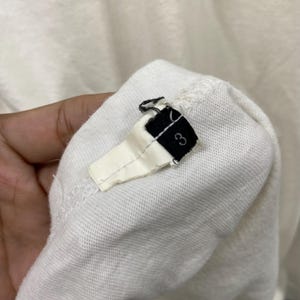 May include: A white fabric garment tag featuring a black rectangular end with the number 3 printed in white. The tag is attached to a white textile surface with visible stitching, showing a folded construction and a frayed edge at the top.