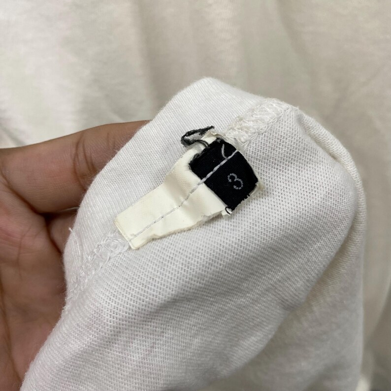 May include: A white fabric garment tag featuring a black rectangular end with the number 3 printed in white. The tag is attached to a white textile surface with visible stitching, showing a folded construction and a frayed edge at the top.