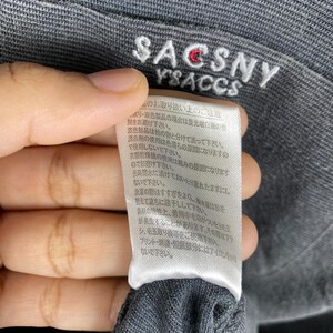 K&ouml;nnte beinhalten: Ein wei&szlig;es Etikett mit schwarzem Text, das Pflegehinweise f&uuml;r ein graues Kleidungsst&uuml;ck enth&auml;lt. Das Etikett lautet "SACSNY YSACCS" und enth&auml;lt Anweisungen zum Waschen, Trocknen und B&uuml;geln des Kleidungsst&uuml;cks.