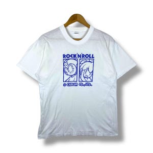 May include: White t-shirt with a blue graphic featuring two cartoon characters and the text "ROCK'N'ROLL". The shirt includes the copyright notice "CAPCOM CO., LTD.". The t-shirt is hanging on a wooden hanger.
