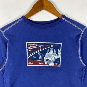 May include: Blue sweatshirt with a white stitched trim around the neck and sleeves. The front of the sweatshirt features a graphic of Buzz Lightyear from the Toy Story franchise. The graphic is a postcard with the text "Greetings from Gamma Quadrant Buzz Lightyear is the Future".