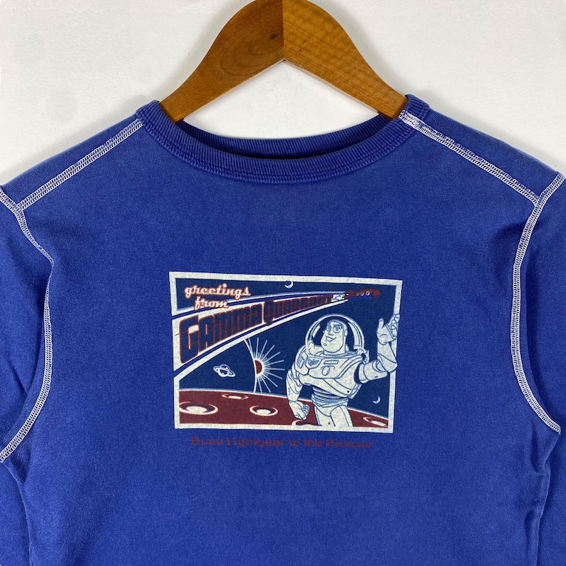 May include: Blue sweatshirt with a white stitched trim around the neck and sleeves. The front of the sweatshirt features a graphic of Buzz Lightyear from the Toy Story franchise. The graphic is a postcard with the text "Greetings from Gamma Quadrant Buzz Lightyear is the Future".