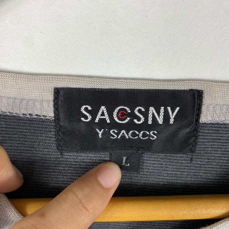 K&ouml;nnte beinhalten: Schwarz-wei&szlig;es Etikett mit dem Text "SACCSNY Y'SACCS" in Wei&szlig;. Das Etikett ist an einem grau-schwarzen Shirt befestigt. Das Gr&ouml;&szlig;enetikett ist sichtbar und zeigt "L".