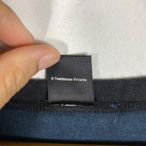 Puede incluir: Una etiqueta negra con el texto "&copy; Touchstone Pictures" en blanco. La etiqueta es rectangular y parece estar cosida a una prenda azul oscuro. La etiqueta est&aacute; sujeta por los dedos de una persona.
