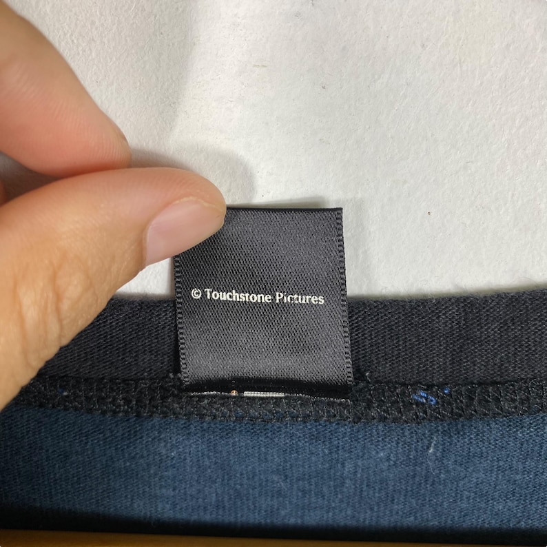 Puede incluir: Una etiqueta negra con el texto "&copy; Touchstone Pictures" en blanco. La etiqueta es rectangular y parece estar cosida a una prenda azul oscuro. La etiqueta est&aacute; sujeta por los dedos de una persona.