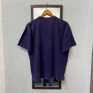 May include: A dark purple t-shirt hanging on a wooden hanger. The shirt is displayed against a mirror with a dark brown frame. The t-shirt has short sleeves and a crew neck.