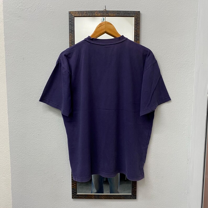May include: A dark purple t-shirt hanging on a wooden hanger. The shirt is displayed against a mirror with a dark brown frame. The t-shirt has short sleeves and a crew neck.