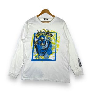 May include: White long-sleeve t-shirt with a graphic print of a cartoon face wearing sunglasses and the word "VOLCOM" in yellow letters. The shirt has a blue and yellow color scheme.