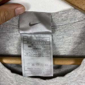 Może przedstawiać: Szara koszulka z białą metką, na kt&oacute;rej widnieje napis "COTTON/POLYESTER FIBERS 4" i "96%/ポリエステル" w języku japońskim. Metka zawiera r&oacute;wnież tekst "(株)ナイキジャパン" i "0120-500-719" w języku japońskim. Metka zawiera r&oacute;wnież napis "MACHINE WASH WARM WITH LIKE COLORS DO NOT BLEACH TUMBLE DRY LOW WARM IRON (DO NOT IRON DESIGN DO NOT DRY CLEAN" i "MADE IN MALAYSIA".