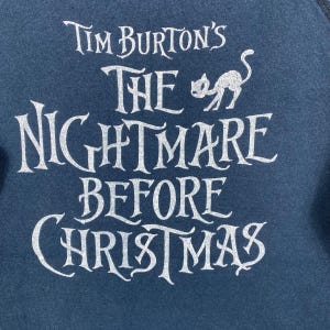 Puede incluir: Sudadera azul oscuro con el texto blanco "Tim Burton's The Nightmare Before Christmas" en letras estilizadas. Una silueta de gato blanco est&aacute; encima de la palabra "Nightmare". Es una prenda de vestir.