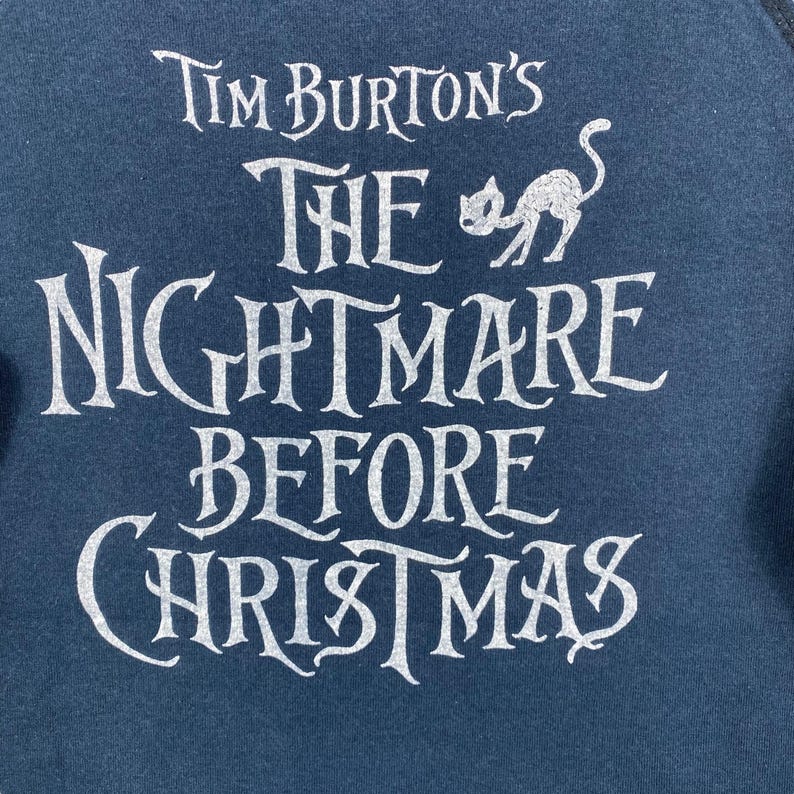 Puede incluir: Sudadera azul oscuro con el texto blanco "Tim Burton's The Nightmare Before Christmas" en letras estilizadas. Una silueta de gato blanco est&aacute; encima de la palabra "Nightmare". Es una prenda de vestir.