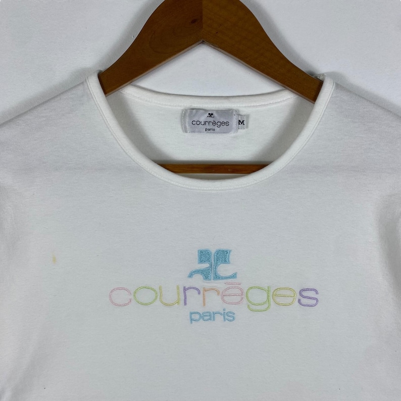May include: White t-shirt with a colorful embroidered logo that says "Courr&egrave;ges Paris". The logo is in a rainbow of colors, including pink, blue, green, and yellow. The shirt has a crew neck and short sleeves. The tag on the shirt says "Courr&egrave;ges Paris" and "M".