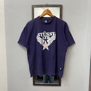 May include: A navy blue t-shirt with the text "ST&Uuml;SSY LTD" in white, featuring a star and wing design. The shirt is hanging on a wooden hanger in front of a mirror.