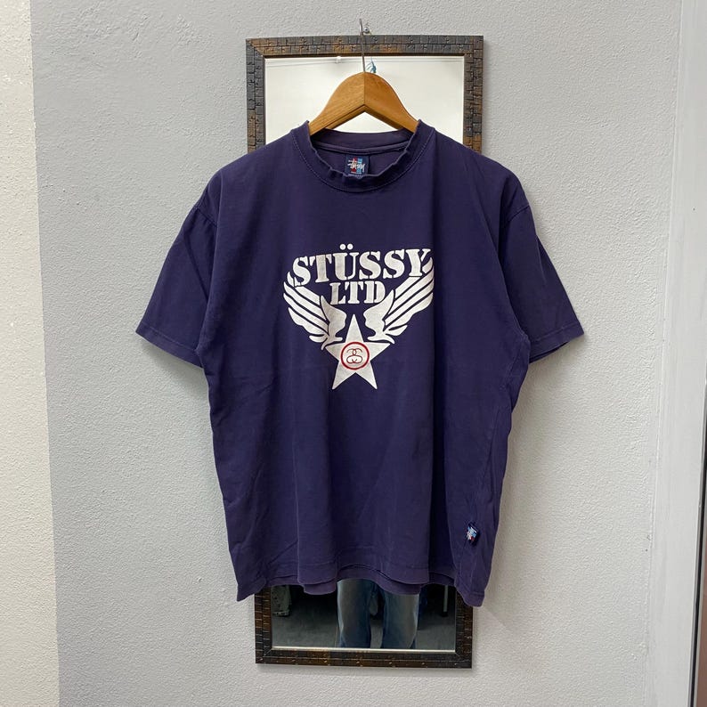 May include: A navy blue t-shirt with the text "ST&Uuml;SSY LTD" in white, featuring a star and wing design. The shirt is hanging on a wooden hanger in front of a mirror.