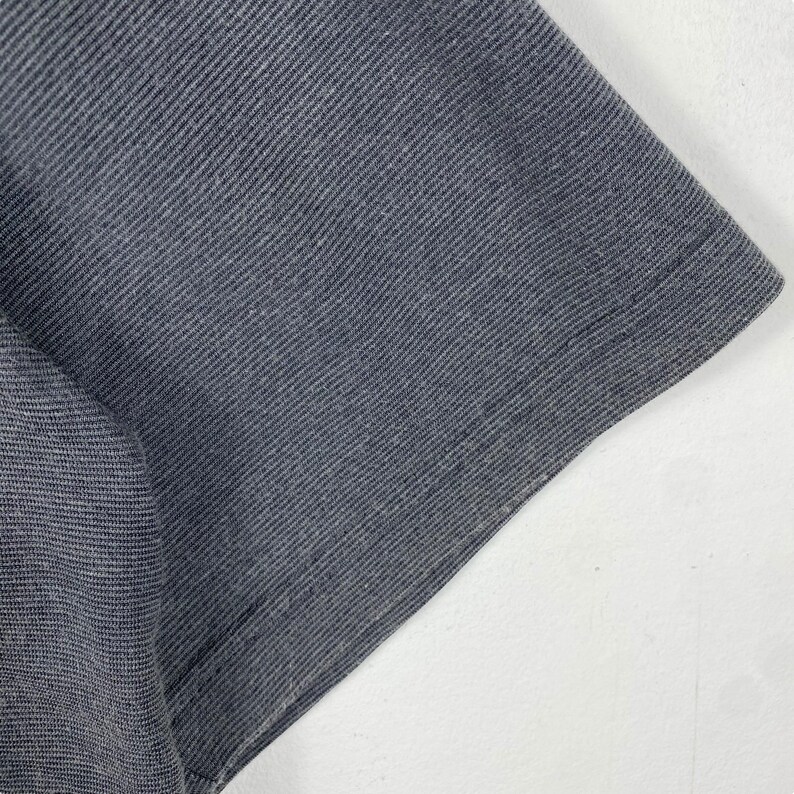 K&ouml;nnte beinhalten: Nahaufnahme des dunkelgrauen Stoffes eines T-Shirts. Der Stoff hat eine strukturierte, leicht gerippte Oberfl&auml;che.