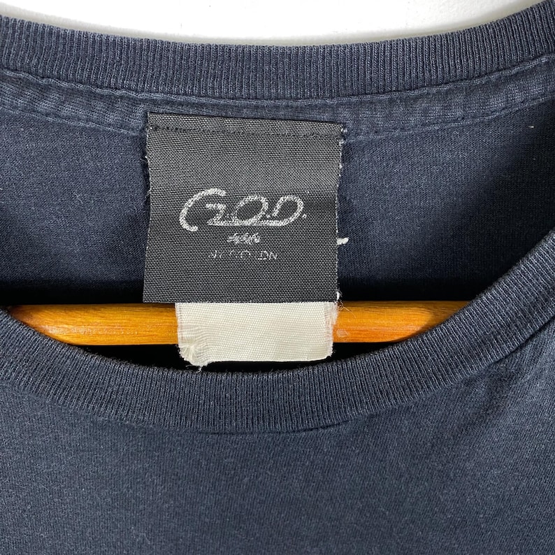 K&ouml;nnte beinhalten: Ein schwarzes T-Shirt mit einem schwarzen Etikett, auf dem "GOD" in wei&szlig;en Buchstaben steht. Das Etikett tr&auml;gt au&szlig;erdem die Aufschrift "J.F.C.O.D.N."