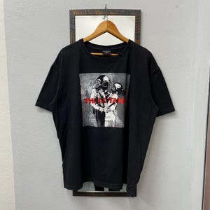 May include: Black t-shirt featuring a graphic of two figures in diving helmets embracing, with the text "THEY TANK" in red. The image is printed on a gray background. The shirt is hanging on a wooden hanger.