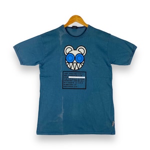 Puede incluir: Una camiseta azul con un gráfico blanco de una criatura de dibujos animados con ojos azules y el texto "TEST SPECIMEN FOR A NEW PHRANACEUTICS HAHE THE COPYRIGHT IN THEIR ORGANISH LIES SOLELY WITH WASTE UN COM A SUBSIDIARY OF RADIOHEAD COM".