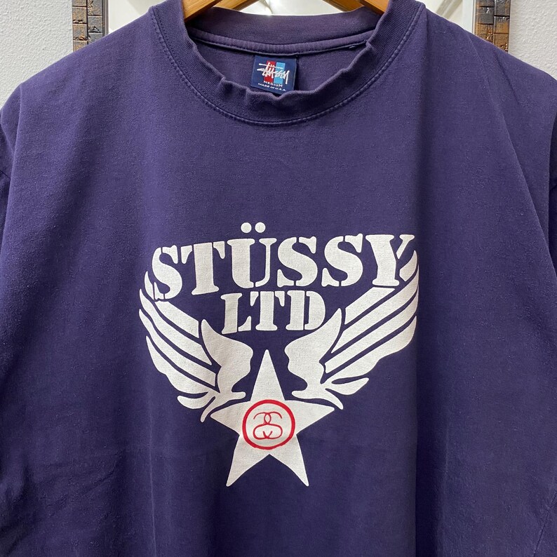 May include: A navy blue St&uuml;ssy LTD t-shirt. The shirt features a large white graphic on the front, including the brand name, wings, and a star with a logo. The shirt has a crew neck and short sleeves.