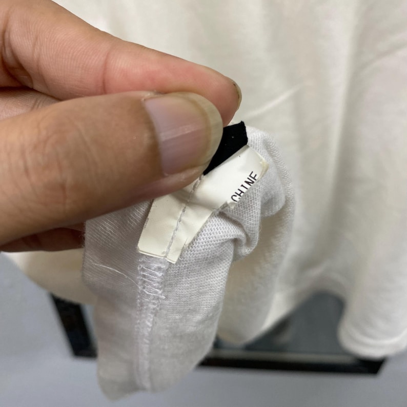 May include: A white fabric garment tag featuring the text CHINE printed in black ink. The tag is attached to a white textile material with visible stitching along the edge, showing the manufacturing details of the clothing item.