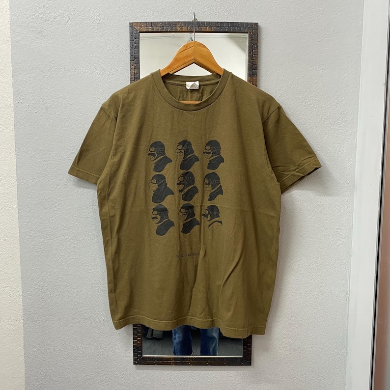 May include: Olive green t-shirt featuring a repeating pattern of black, stylized faces with goggles. The shirt is hanging on a wooden hanger in front of a mirror. The text "HARDCORE" is printed at the bottom of the shirt.