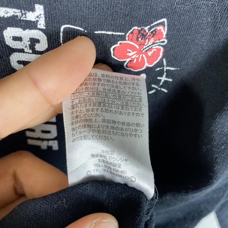 Pode incluir: Camiseta preta com um gr&aacute;fico branco e um design floral vermelho. A camisa tem texto japon&ecirc;s e uma etiqueta com mais texto japon&ecirc;s. A etiqueta tamb&eacute;m tem o texto "SANRIO".