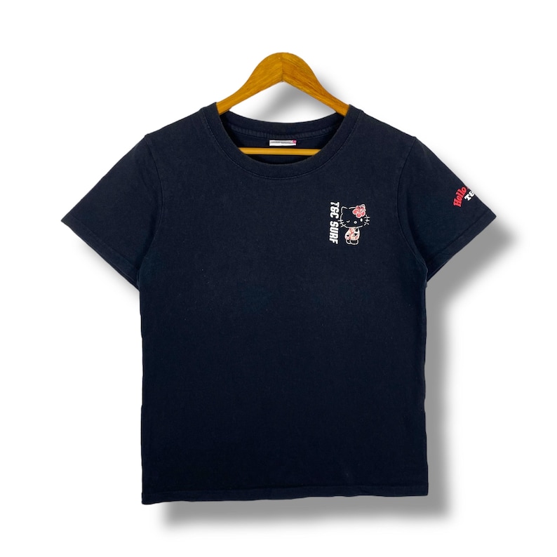 Pode incluir: Camiseta preta de gola redonda com um pequeno gr&aacute;fico de um gato usando uma flor e o texto "TSC SURF" na frente. A manga tem o texto "Hello Kitty". A camisa est&aacute; pendurada em um cabide de madeira.
