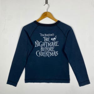Puede incluir: Camiseta de manga larga azul marino con el texto blanco "Tim Burton's The Nightmare Before Christmas". La camiseta tiene un cuello negro y mangas ragl&aacute;n, y est&aacute; colgada en una percha de madera.