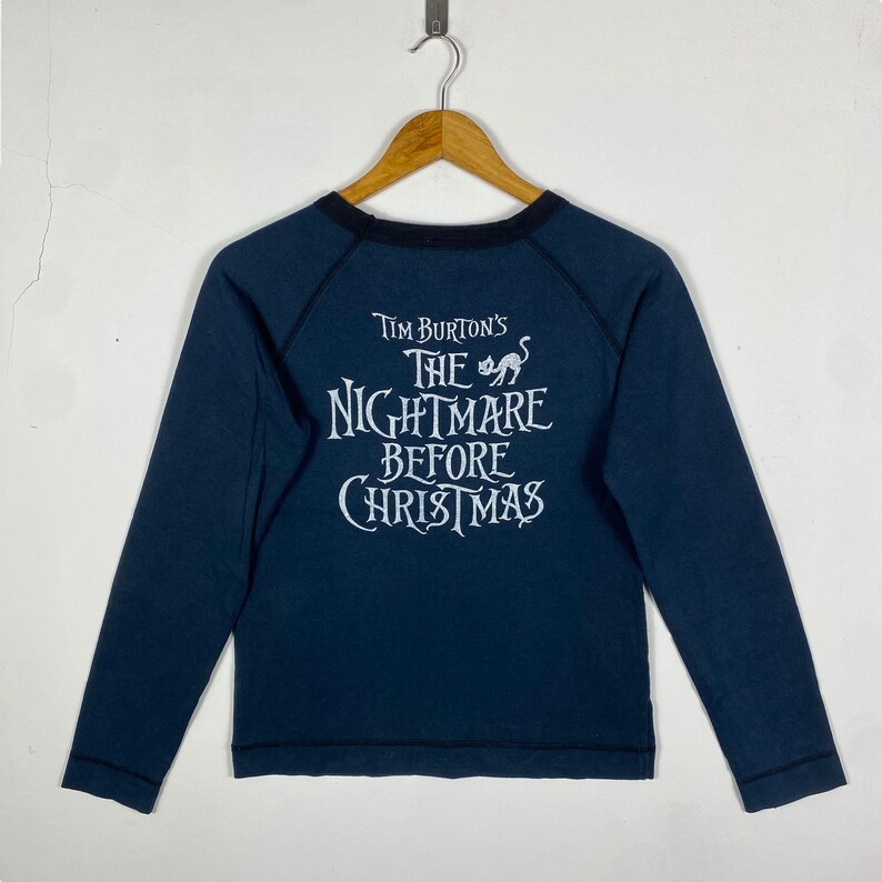Puede incluir: Camiseta de manga larga azul marino con el texto blanco "Tim Burton's The Nightmare Before Christmas". La camiseta tiene un cuello negro y mangas ragl&aacute;n, y est&aacute; colgada en una percha de madera.
