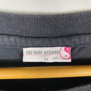 Pode incluir: Close-up da gola de uma camiseta preta com uma etiqueta. A etiqueta diz "TGC SURF DESIGNS" em texto rosa, com um s&iacute;mbolo yin-yang. O tamanho "LL" est&aacute; impresso abaixo. A camisa est&aacute; pendurada em um cabide de madeira.