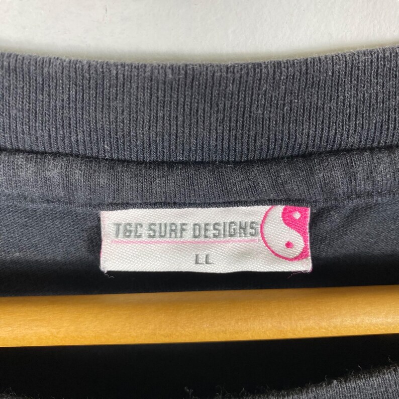 Pode incluir: Close-up da gola de uma camiseta preta com uma etiqueta. A etiqueta diz "TGC SURF DESIGNS" em texto rosa, com um s&iacute;mbolo yin-yang. O tamanho "LL" est&aacute; impresso abaixo. A camisa est&aacute; pendurada em um cabide de madeira.