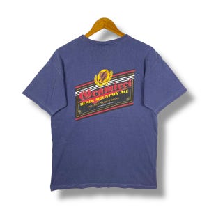 Peut inclure: Un t-shirt bleu avec un graphisme au dos. Le graphisme est une étiquette rectangulaire avec les mots "Gramicci Black Mountain Ale" dans une police stylisée. L'étiquette a des accents noirs, rouges et dorés.