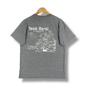 May include: Gray short-sleeved t-shirt with a white graphic print of a group of people on a vehicle. The text "Team Darai" and "ガールズ＆パンツァー" are printed on the shirt.