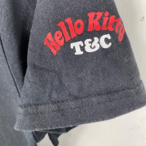 Pode incluir: Close-up de uma manga de camiseta cinza escuro. A manga apresenta o texto "Hello Kitty" em vermelho, com "T&C" em branco abaixo. O tecido tem uma apar&ecirc;ncia ligeiramente desgastada.