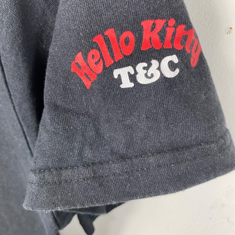 Pode incluir: Close-up de uma manga de camiseta cinza escuro. A manga apresenta o texto "Hello Kitty" em vermelho, com "T&C" em branco abaixo. O tecido tem uma apar&ecirc;ncia ligeiramente desgastada.