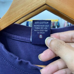May include: Close-up of a navy blue t-shirt tag. The tag reads "100% COTTON, MACHINE WASH COLD, TUMBLE DRY LOW, HAVE A GOOD VIBE! CA-28629". The tag also features a logo. The t-shirt is made of cotton.