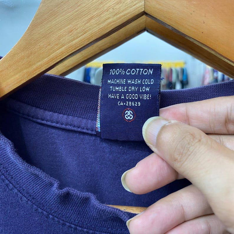 May include: Close-up of a navy blue t-shirt tag. The tag reads "100% COTTON, MACHINE WASH COLD, TUMBLE DRY LOW, HAVE A GOOD VIBE! CA-28629". The tag also features a logo. The t-shirt is made of cotton.
