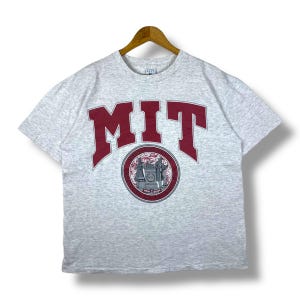 Może przedstawiać: Szara koszulka z krótkim rękawem z czerwonym logo "MIT" i okrągłym nadrukiem pieczęci Massachusetts Institute of Technology.