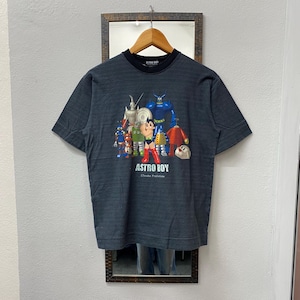 Peut inclure: T-shirt gris foncé avec un motif coloré d'Astro Boy et d'autres personnages. Le t-shirt a un col noir et des manches courtes. Le texte "ASTRO BOY" est imprimé en dessous. Un t-shirt de style vintage.