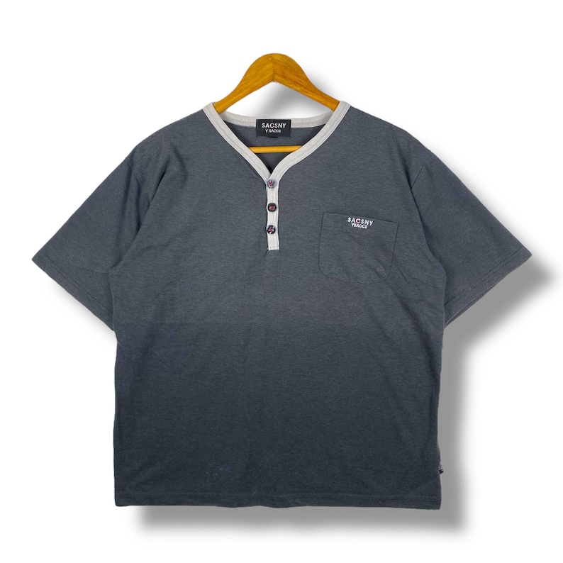 K&ouml;nnte beinhalten: Ein graues T-Shirt mit kurzen &Auml;rmeln, einem wei&szlig;en Kragenbesatz und einer Knopfleiste. Das Shirt hat eine kleine Tasche auf der linken Brust mit dem gestickten Schriftzug "SACSINY FRANCE". Der Schriftzug "SACSINY FRANCE" ist auch am Nacken gestickt.