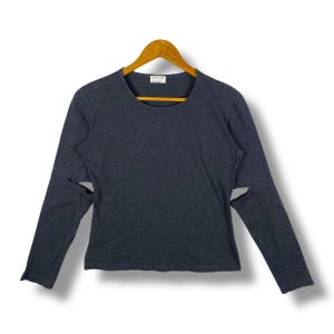 Puede incluir: Una camiseta de manga larga gris oscuro con cuello redondo y detalles recortados en las mangas. La camiseta se muestra en una percha de madera sobre un fondo blanco. La camiseta parece estar hecha de un material suave y cómodo.