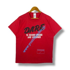 Puede incluir: Camiseta roja de manga corta con el texto "DARE TO RESIST DRUGS AND VIOLENCE" en letras negras. La camiseta también tiene una pequeña etiqueta rectangular con texto.