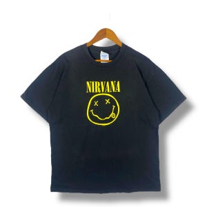 Może przedstawiać: Czarny T-shirt z żółtą uśmiechniętą buźką i nadrukiem "NIRVANA" z przodu.