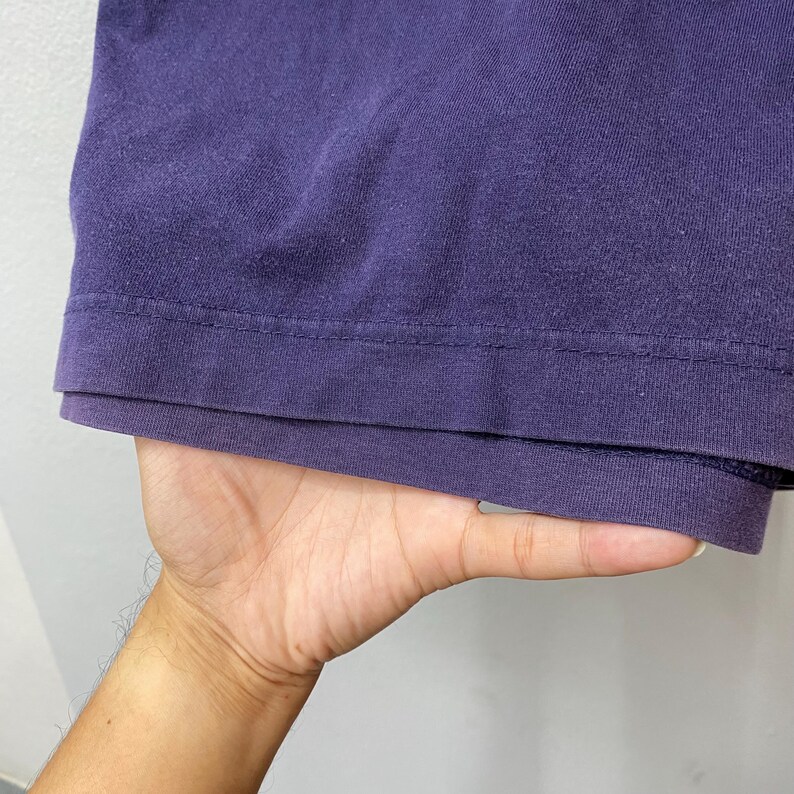 May include: Close-up of a deep purple t-shirt hem. The fabric appears soft and slightly worn, with a visible seam detail. The shirt is held by a hand, suggesting it's being displayed or examined. The overall impression is of a casual, everyday garment.