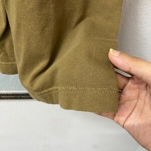 May include: Close-up of a section of a brown t-shirt. The fabric is a solid color with visible texture and a stitched hem. The image shows the edge of the shirt and a hand touching the fabric.
